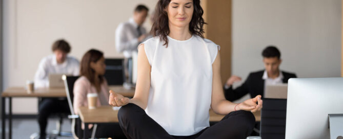 Frau macht Yogaübung im Büro auf dem Schreibtisch während andere Mitarbeiter:innen weiter arbeiten.